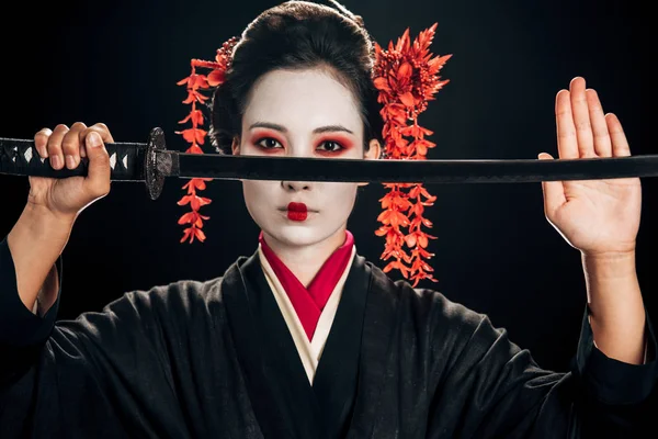 Geisha confiada en kimono negro con flores rojas en el pelo sosteniendo katana aislado en negro - foto de stock