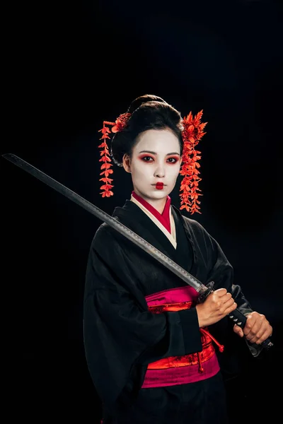 Schöne Geisha im schwarzen Kimono mit roten Blumen im Haar, die Katana isoliert auf Schwarz hält — Stockfoto