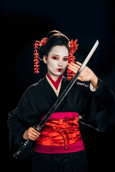 Belle geisha en kimono noir avec des fleurs dans les cheveux tenant katana isolé sur noir — Photo de stock