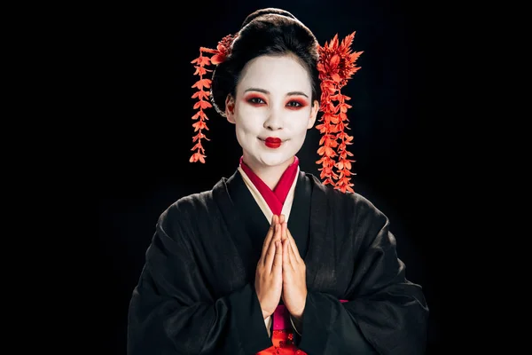 Side view of happy beautiful geisha in black and red kimono and flowers in hair looking at camera and greeting isolated on black — Stock Photo