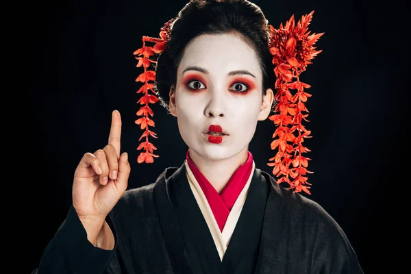 Schockiert schöne Geisha in schwarz-rotem Kimono und Blumen im Haar, die Ideengeste isoliert auf schwarz zeigen — Stockfoto