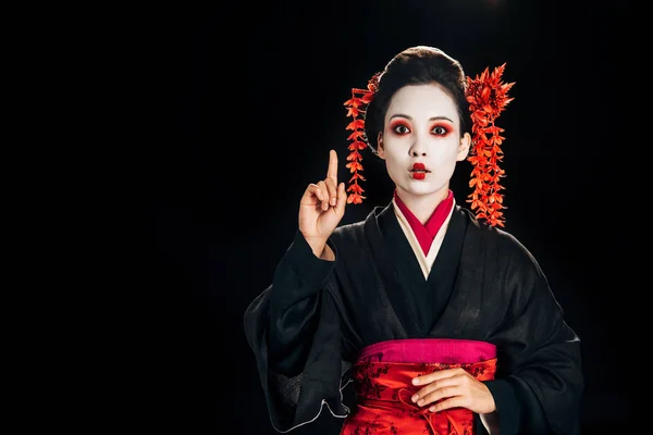 Shocked beautiful geisha in black and red kimono and flowers in hair showing idea gesture isolated on black with copy space — Stock Photo