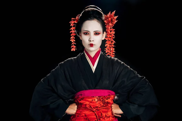 Geisha hermosa enojada en kimono negro y rojo y flores en el pelo con las manos en las caderas aisladas en negro - foto de stock
