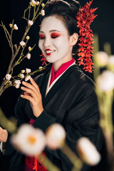 Selective focus of happy beautiful geisha in black kimono with red flowers in hair and sakura branches isolated on black — Stock Photo