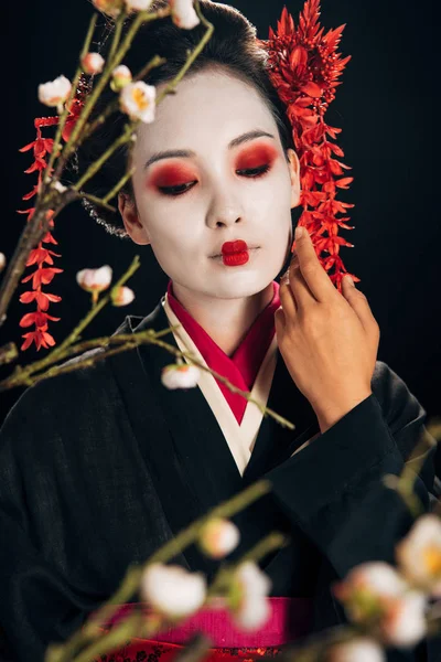 Selective focus of beautiful geisha in black kimono with red flowers in hair touching face and sakura branches isolated on black — Stock Photo