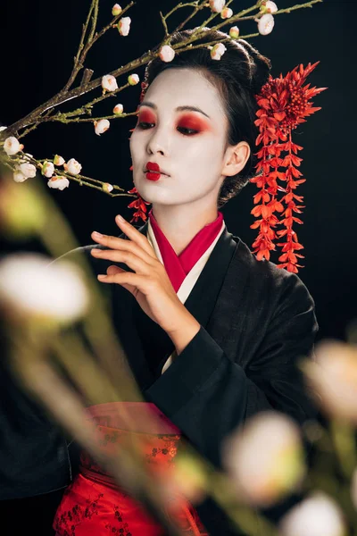 Selektiver Fokus der schönen Geisha im schwarzen Kimono mit roten Blüten im Haar und Sakura-Zweigen auf Schwarz — Stockfoto