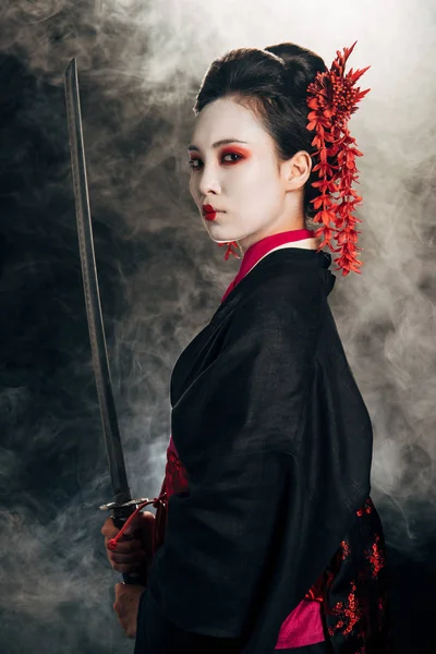 Side view of beautiful geisha in black kimono holding katana in smoke on black background — Stock Photo