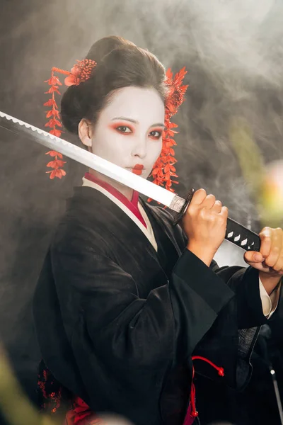 Селективний фокус гейші в чорному кімоно тримає катану в диму і гілках сакури — стокове фото