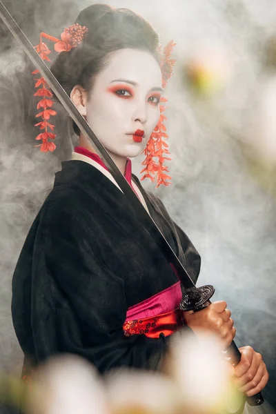 Foco selectivo de geisha seria en kimono negro sosteniendo katana en ramas de humo y sakura - foto de stock