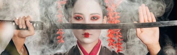 Selective focus of beautiful geisha in black kimono holding katana in smoke, panoramic shot — Stock Photo