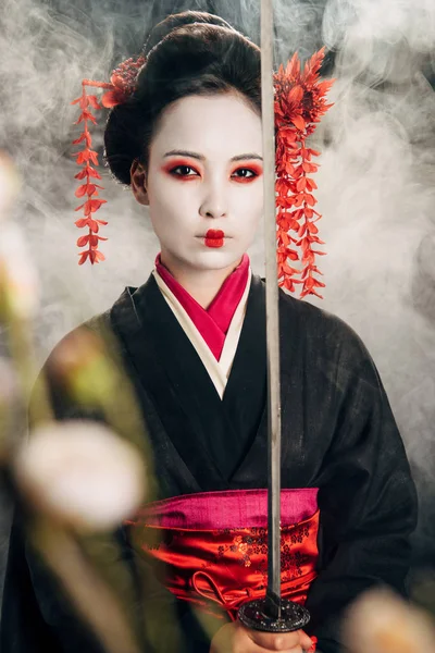 Foyer sélectif de belle geisha en kimono noir tenant katana en fumée et branches de sakura — Photo de stock