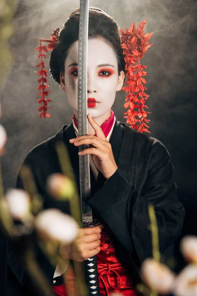 Селективный фокус гейши в черном кимоно, держащей катану в дыму и ветвях сакуры — стоковое фото