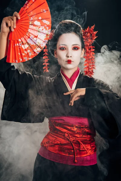 Beautiful geisha in black kimono with flowers in hair holding hand fan and gesturing in smoke — Stock Photo