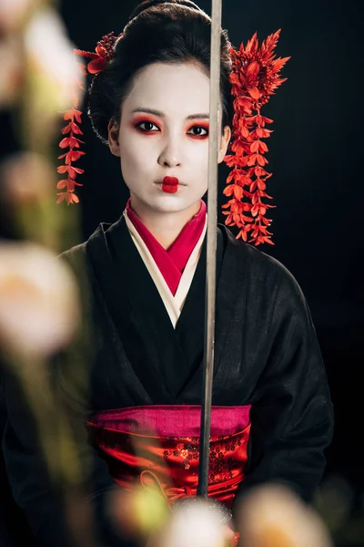 Selective focus of confident geisha in black kimono holding katana and sakura branches isolated on black — Stock Photo