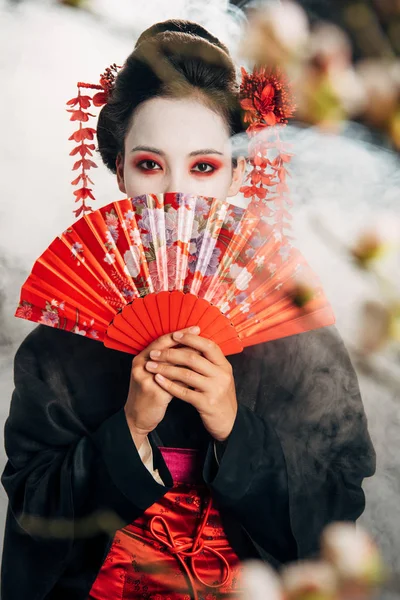 Foco seletivo da bela gueixa em quimono preto com flores no cabelo segurando ventilador de mão na frente do rosto e ramos sakura em fumaça — Fotografia de Stock