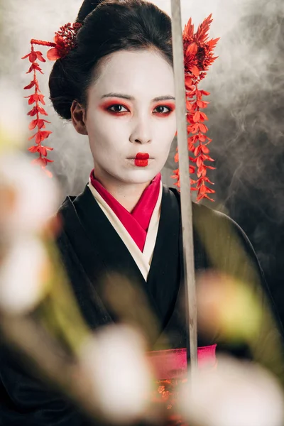 Selective focus of confident geisha in black kimono holding katana in smoke and sakura branches — Stock Photo