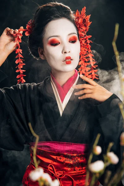 Foco seletivo da bela gueixa em quimono preto com flores vermelhas no cabelo e ramos sakura no fundo preto com fumaça — Fotografia de Stock