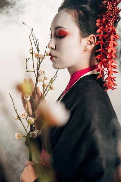 Selective focus of beautiful geisha in black kimono with red flowers in hair and sakura branches in smoke — Stock Photo