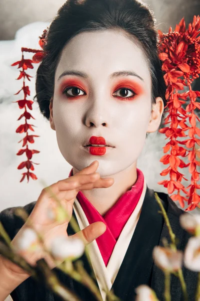Portrait of beautiful geisha in black kimono with red flowers in hair and sakura branches on black background with smoke — Stock Photo