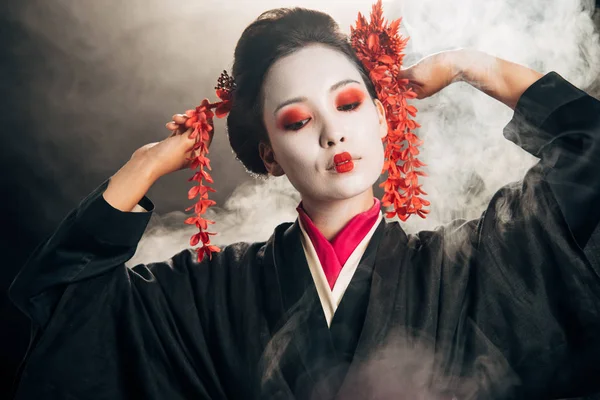 Schöne Geisha im schwarzen Kimono mit roten Blumen im Haar gestikulierend auf schwarzem Hintergrund mit Rauch — Stockfoto