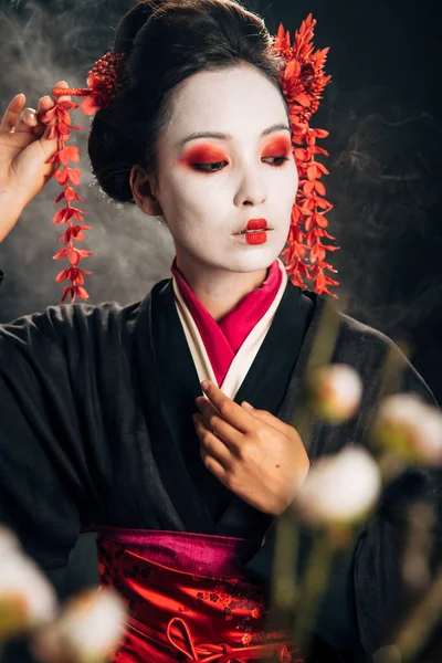 Foco seletivo da gueixa em quimono preto com flores vermelhas no cabelo e ramos sakura em fundo preto com fumaça — Fotografia de Stock