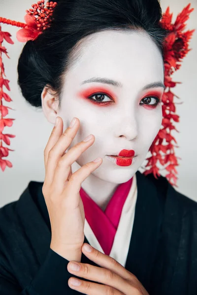 Portrait of beautiful geisha in black kimono with red flowers in hair touching face isolated on white — Stock Photo