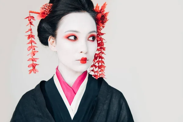Hermosa geisha en kimono negro con flores rojas en el pelo mirando hacia otro lado aislado en blanco - foto de stock