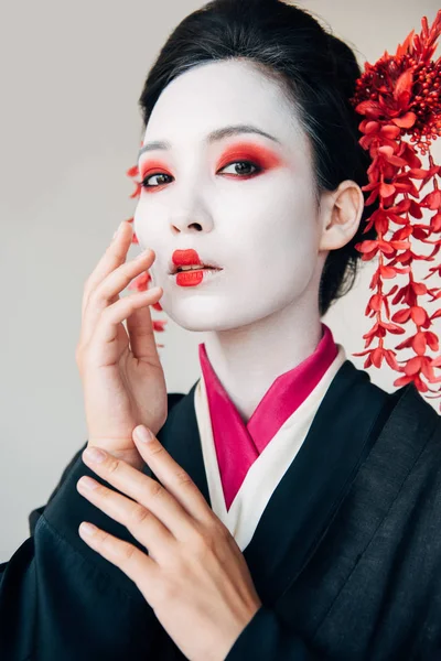 Schöne Geisha im schwarzen Kimono mit roten Blumen im Haar, die Gesicht berühren, isoliert auf weiß — Stockfoto