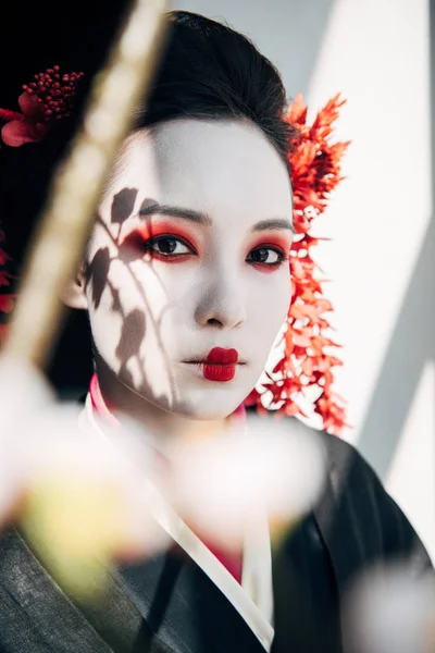 Selective focus of sakura branches and beautiful geisha with red and white makeup in sunlight — Stock Photo