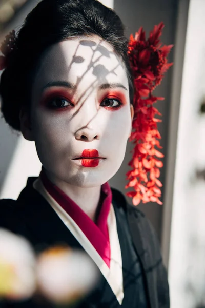 Selective focus of sakura branches and beautiful geisha with red and white makeup in sunlight — Stock Photo