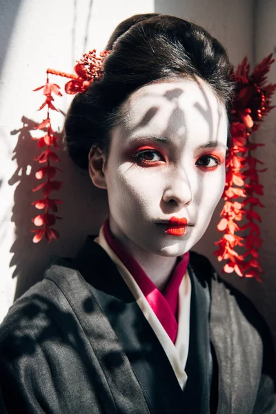 Portrait of beautiful geisha with red and white makeup looking at camera in sunlight with shadows — Stock Photo