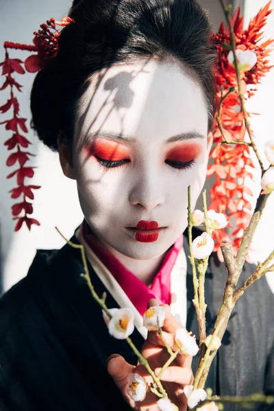 Ramas de sakura y hermosa geisha con maquillaje rojo y blanco a la luz del sol - foto de stock