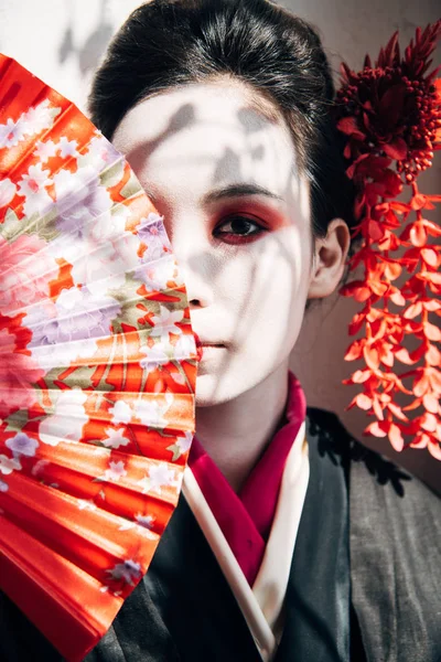 Hermosa geisha con maquillaje rojo y blanco sosteniendo ventilador de la mano cerca de la cara a la luz del sol - foto de stock