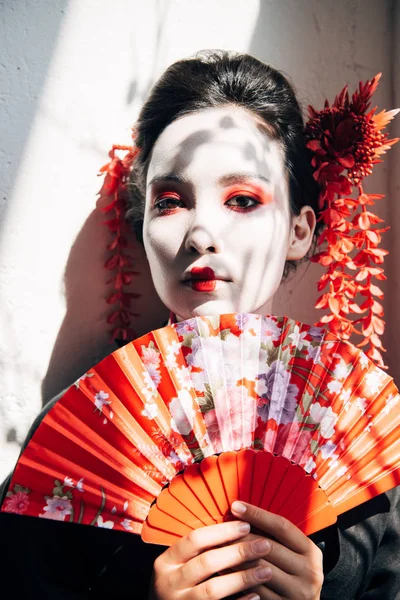 Portrait of beautiful geisha with red and white makeup holding hand fan in sunlight — Stock Photo