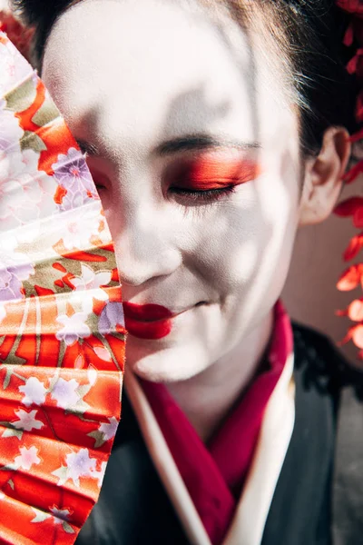 Портрет усміхненої красивої гейші з червоно-білим макіяжем, що тримає вентилятор рук на сонячному світлі — стокове фото