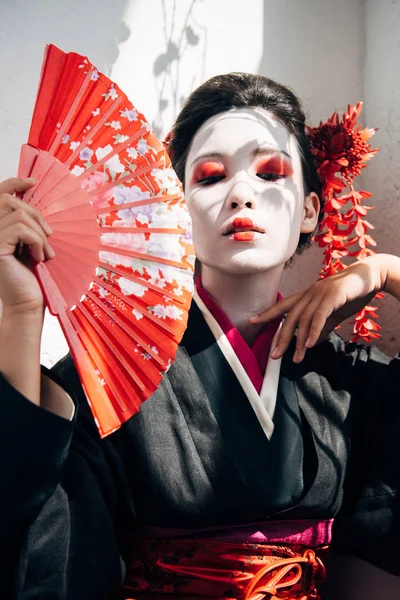 Portrait of beautiful geisha with red and white makeup holding hand fan and gesturing in sunlight — Stock Photo