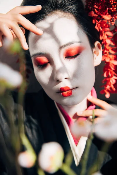Вибірковий фокус гілок дерева і красива гейша з червоно-білим макіяжем і закритими очима на сонячному світлі — стокове фото