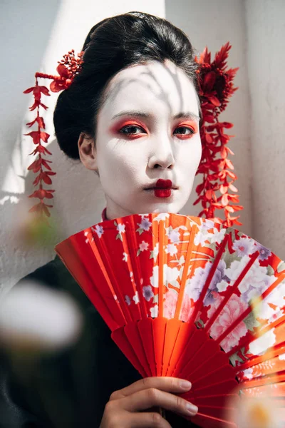 Messa a fuoco selettiva di rami d'albero e bella geisha con trucco rosso e bianco tenendo ventilatore a mano alla luce del sole — Foto stock