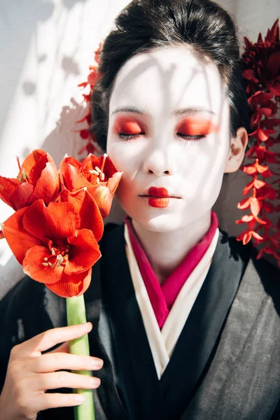 Portrait of beautiful geisha holding red flowers in sunlight — Stock Photo
