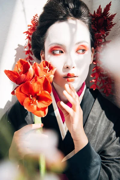 Selective focus of sakura branches and beautiful geisha holding red flowers in sunlight — Stock Photo