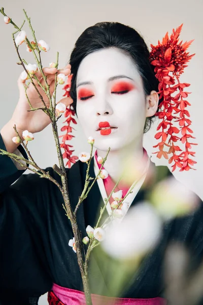 Selektiver Fokus von Baumzweigen und wunderschöner Geisha mit rot-weißem Make-up und geschlossenen Augen isoliert auf weiß — Stockfoto