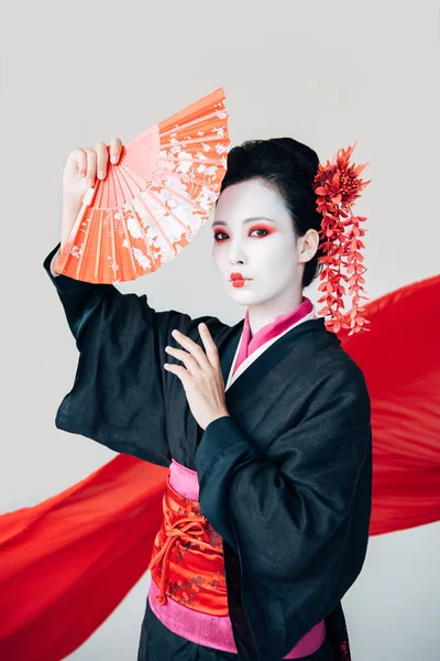 Hermosa geisha en kimono negro posando con ventilador de mano y tela roja sobre fondo aislado en blanco - foto de stock