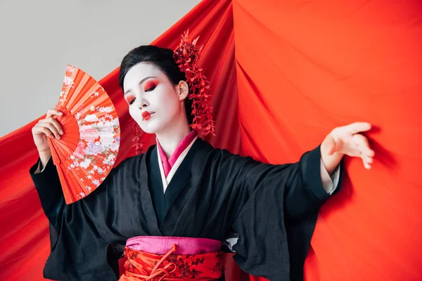 Hermosa geisha en kimono negro con abanico de mano y tela roja sobre fondo bailando aislado en blanco - foto de stock