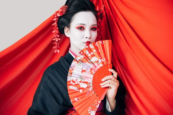 Beautiful geisha in black kimono with hand fan and red cloth on background isolated on white — Stock Photo