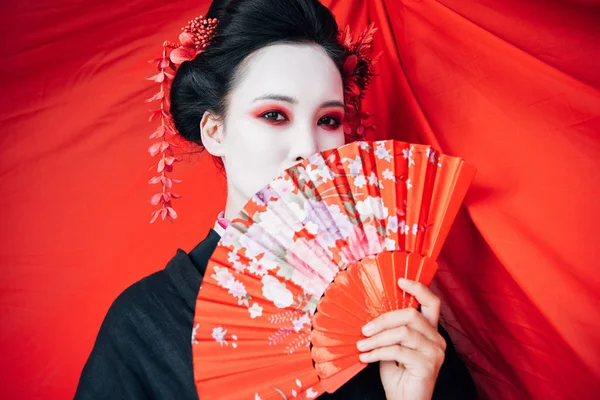 Beautiful geisha in black kimono with hand fan and red cloth on background — Stock Photo