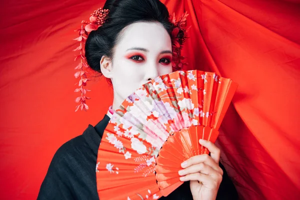 Schöne Geisha im schwarzen Kimono mit Handfächer und rotem Tuch auf Hintergrund — Stockfoto