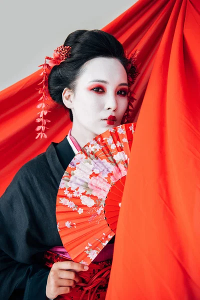 Hermosa geisha en kimono negro con ventilador de mano cerca de tela roja sobre fondo aislado en blanco - foto de stock