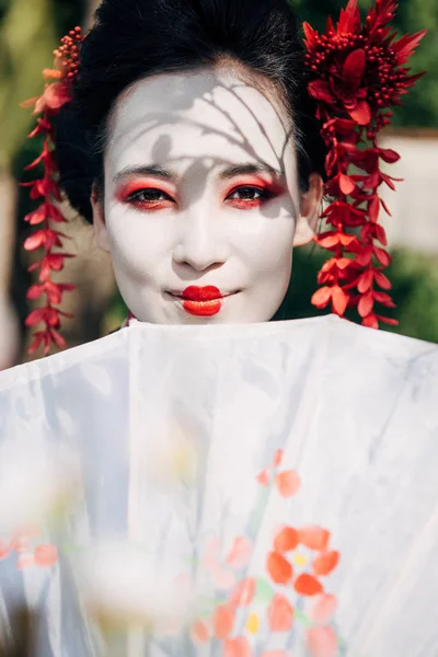 Selective focus of tree branches and beautiful smiling geisha with umbrella in sunlight — Stock Photo