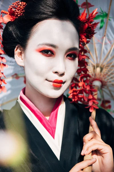 Selective focus of beautiful smiling geisha with umbrella in sunlight — Stock Photo