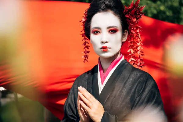 Selective focus of tree branches and beautiful geisha with greeting gesture looking at camera with red cloth on background in sunlight — Stock Photo